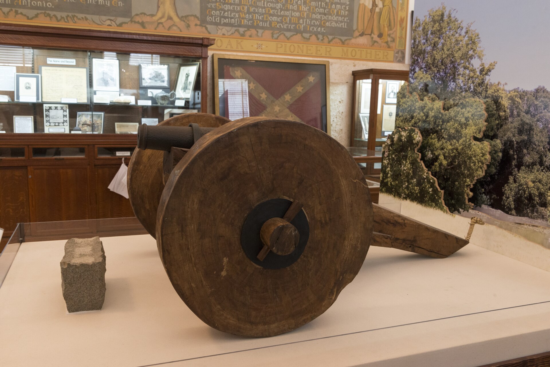 A small cannon on solid wooden wheels in a glass case at the Gonzales Memorial Museum