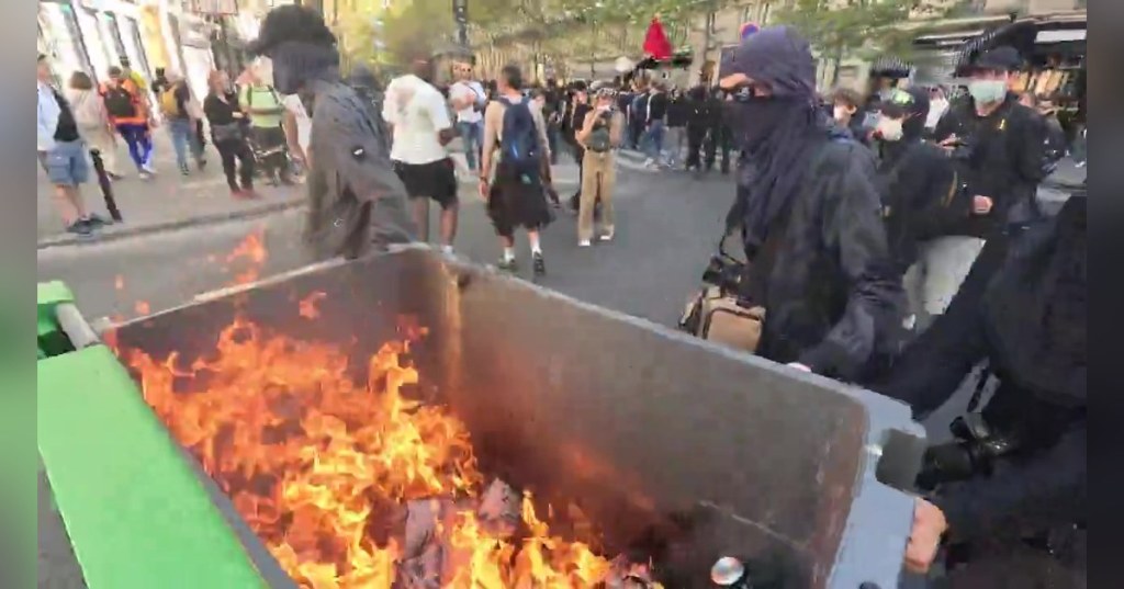Frame from the video posted by @nickshirleyy on X: masked protesters beside a burning trash container on a Paris street